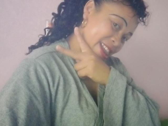 Rahel69 with curly black hair, light brown skin, and gold hoop earrings, wearing a gray shirt, making a peace sign near her chin, smiling with pink lipstick.