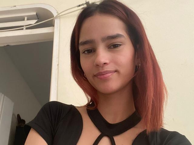 MelodyMansson has red-brown hair, a small nose, and a subtle smile. She's wearing a black top with cut-out neck, showing her tits. She's a cute, sexy girl.