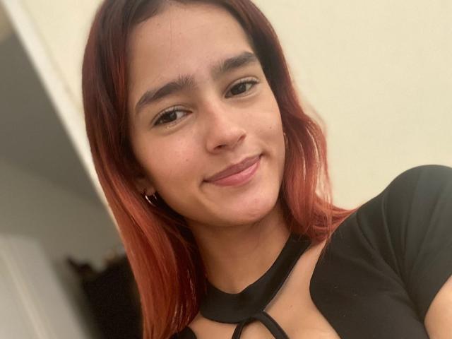 MelodyMansson has red-tipped hair, dark eyebrows, and a slight smile, wearing a black top with a keyhole cutout, showing a bit of her chest.