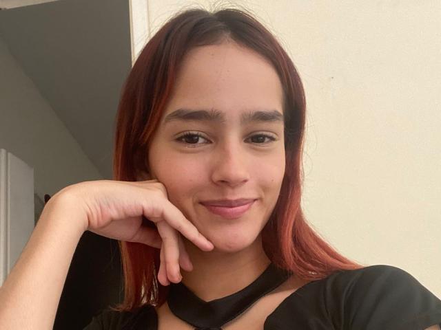 MelodyMansson with red-tinted hair, medium skin, and a subtle smile, resting her chin on her hand, wearing a black top with a choker, in a casual selfie.