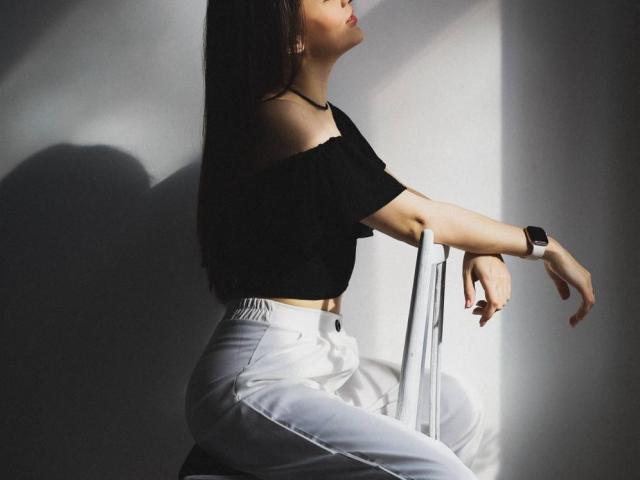 TenerliRize, with medium tits, wearing a black off-shoulder top and white pants, sits on a white chair in sunlight, shadow on her left, wristwatch on her right.