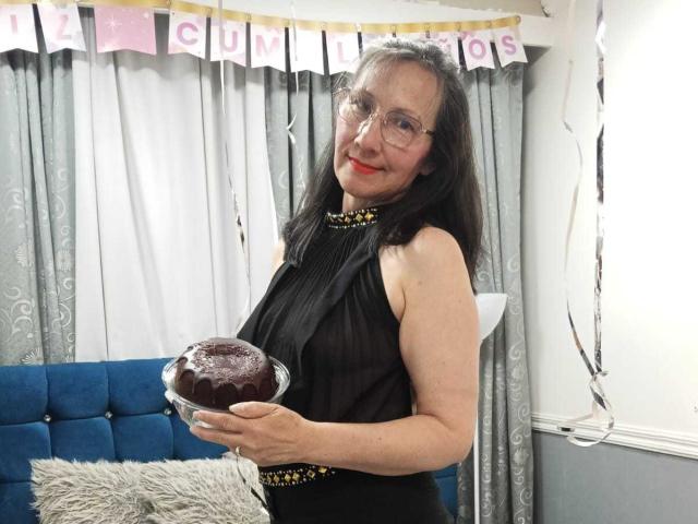 OrianaJhonson with glasses, black sleeveless dress, holding glass of dark chocolate pie, smiling, red lipstick, gray curtains, blue couch, white floor lamp, gray wall, white trim.