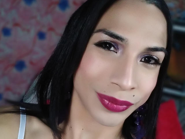 BigCockLatinxx with black hair, purple eyeshadow, thick eyebrows, pink lipstick, and a small beauty mark on her right cheek, smiling at the camera.