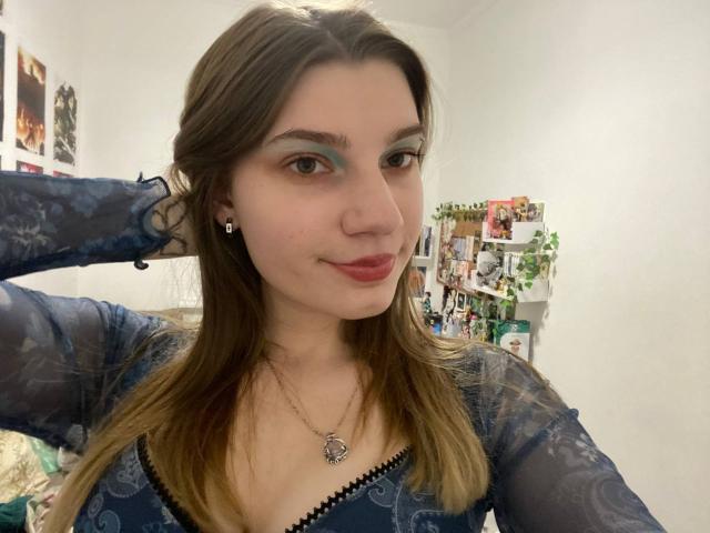 PureRadiance with brown hair, green eye makeup, and blue sheer top, showing off her tits with a hand in her hair, wearing a heart necklace, smiling softly.