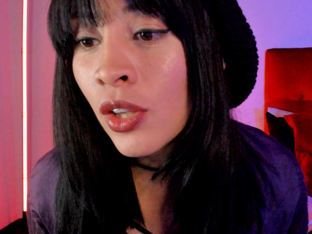 IssisDiosa with dark straight hair, black beanie, black choker, and glossy red lipstick, looking slightly to the side with a subtle smile, wearing a black shirt.