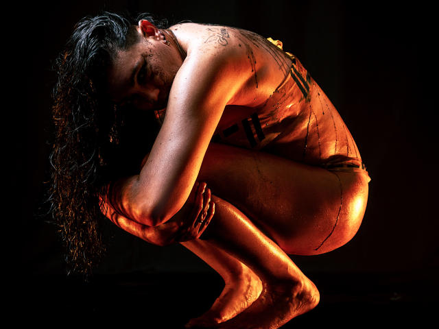 SaharaMK is crouching, naked, with wet, black-tattooed skin, and curly dark hair. Her ass is highlighted by orange lighting, and she's gripping her ankle.