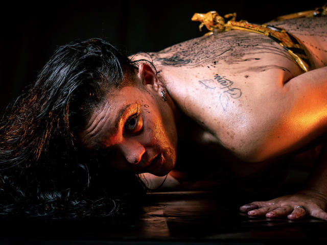 SaharaMK is on all fours, dirty and sweaty, with dark wet hair, golden eyes, tattooed shoulder, and a gold ornament on their back. Their face is close to the ground.