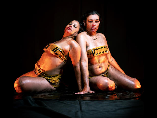 SaharaMK with tan skin, dark hair, and wet body paint sits on black cloth, back to back with SaharaMK, wearing gold band-tied tops and bottoms, looking at camera.