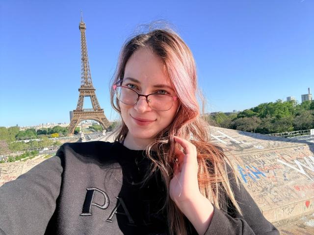 AmbeerWood with pinkish-brown hair, glasses, and black sweater, holding hair, smiling, standing in front of the Eiffel Tower, clear blue sky, sunny, green trees in background.