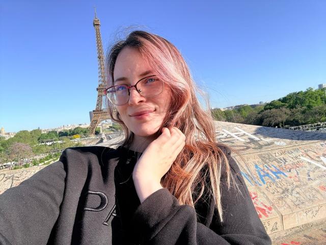 AmbeerWood with pinkish-brown hair, glasses, and black coat, smiling, hand on neck, Eiffel Tower in background, clear blue sky, graffiti-covered wall, sunlight, casual, cute.