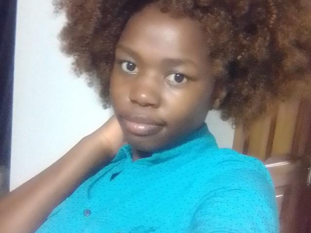 Yollah has dark brown skin, curly brown afro, and is wearing a teal button-up shirt. She's looking at the camera with a slight smile, hand near her neck.