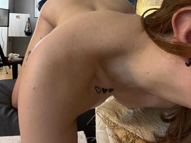 VictoriyaOrtega's bare back and ass are visible, with small black heart tattoos on her side and a brown-haired side profile. She's bent over, showing her arched back.