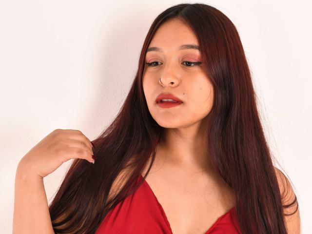 MarianaVargasX with long red hair, red lipstick, and pink eyeshadow, wearing a red dress, touching her hair with her right hand, showing a slight pout.