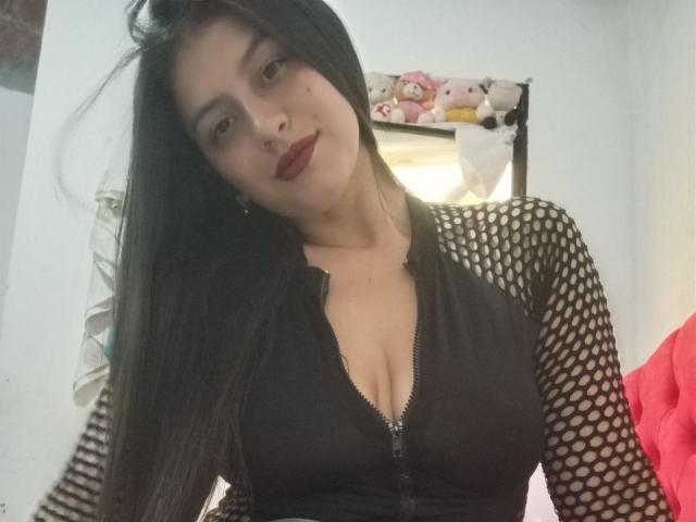 MadissonFontaine with long black hair, wearing a black fishnet top with a deep cleavage zipper, red lipstick, and a slight smile, in a bedroom with stuffed animals.