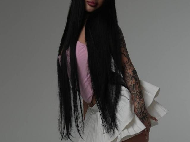 ChristieHoliday with long black hair, pink top, white pleated skirt, and tattooed arm, showing her ass slightly. Her tattoos are on her right arm.