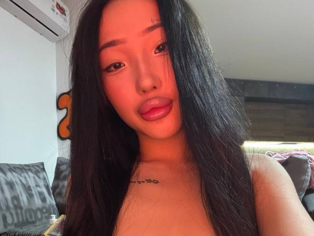 ChristieHoliday with long black hair, red-tinted skin, glossy pink lips, and a small tattoo under her chin, showing her bare shoulders and upper chest.