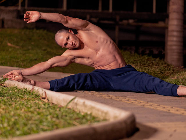 AxelFairchail is shirtless, muscular, and doing a split stretch on a sidewalk at night, wearing blue pants, with a raised arm and focused expression. His ass is toned.