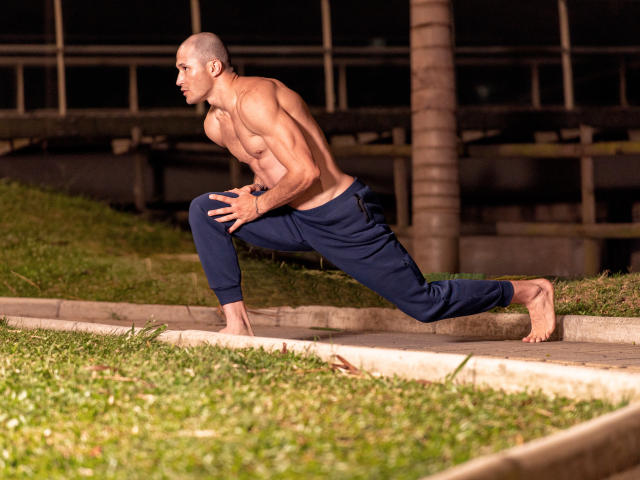 AxelFairchail is shirtless, muscular, bald, and doing a lunge on grass at night. He's wearing dark blue pants, barefoot, with a focused expression.