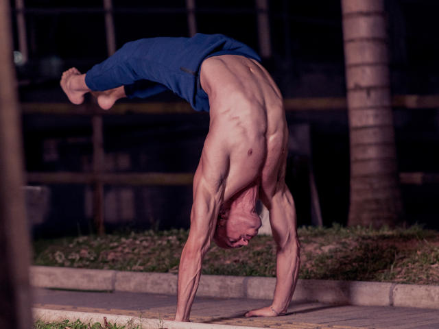 AxelFairchail, shirtless with muscular back and arms, doing a headstand, wearing blue pants, outside at night, with a palm tree in the background.