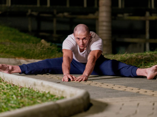 AxelFairchail, bald, muscular, white t-shirt, blue pants, split in a crouched position, hands on pavement, bare feet, focused expression, night setting, grass, and a concrete edge in foreground.