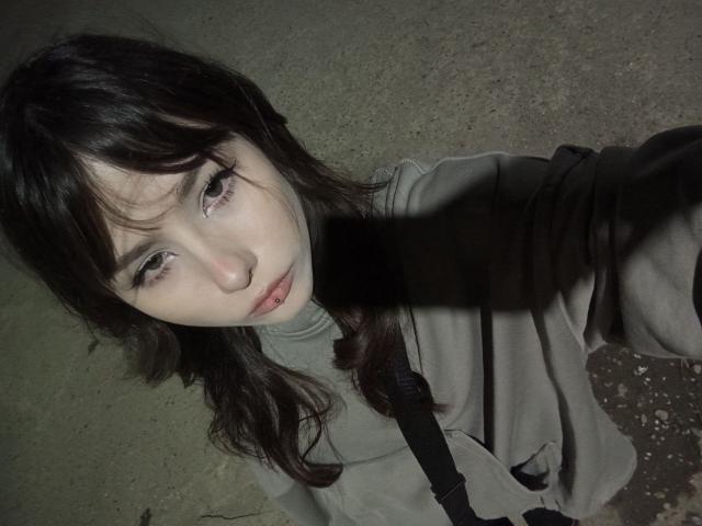 EileneSzala with dark, wavy hair, pale skin, and sad eyes, wearing a gray hoodie, has a black shadow across her face, sitting on a rough, textured surface.