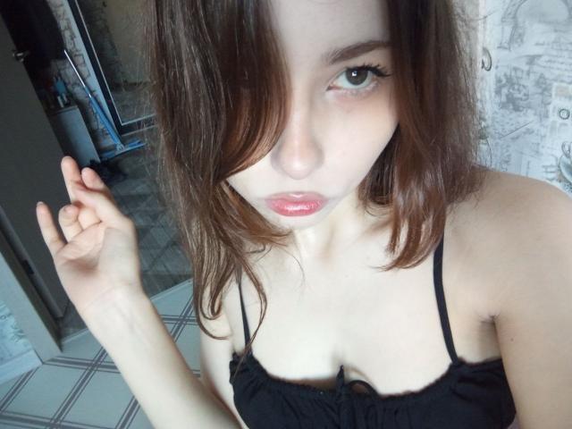 EileneSzala with brown hair, green eyes, and pink lips, wearing a black spaghetti-strap top, showing small tits and pale skin, raising her left hand.