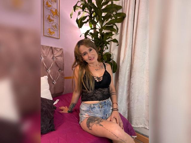 SharoLoveDion with long blonde hair, black lace top, and denim shorts, sitting on a pink bed, showing her tattoos and smiling. She's got small boobs and a cute face.