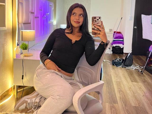NyraVanel with black top, grey pants, sitting in white chair, holding phone, smiling, wooden floor, lamp, mirror, phone case with cow pattern, black hair, small tattoo on chest.