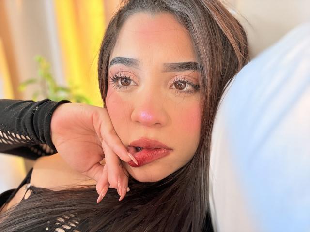 LunaVali with red lipstick, dark hair, and long eyelashes, resting her hand on her chin, wearing a black mesh sleeve, looking at the camera with a slight smile.