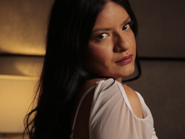 SophieSoler with long black hair, brown eyes, and pink lipstick, wearing a white sleeveless top, looking over her shoulder with a slight smile, showing her bare shoulder.