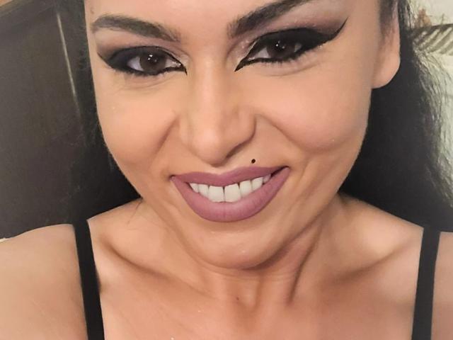 ExquisDunya has dark, dramatic eye makeup, a small mole on her upper lip, and is smiling with pink lipstick, wearing a black strap top, showing her tits.