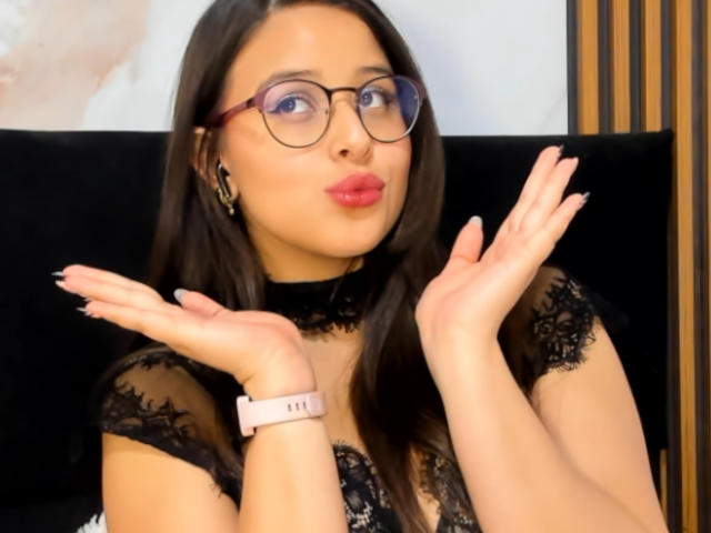 SunnyLuxx with pouty lips, wearing glasses, black lace top, pink watch, and earrings, hands up, looking cute. Brown hair, light skin, red lipstick.