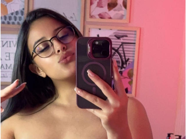 SunnyLuxx with glasses, taking a mirror selfie, holding a phone with a ring design, showing her bare shoulders and pouty lips, pink light in the background.