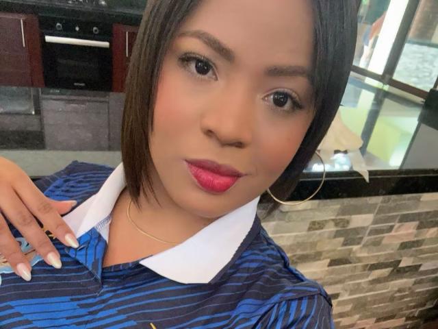 AliceBooty with short brown hair, red lipstick, blue striped polo with white collar, gold necklace, and hoop earrings, showing off manicured nails in a kitchen setting.