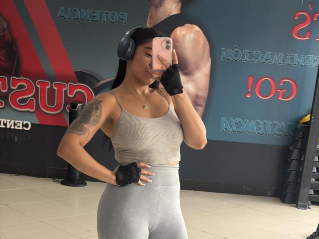StellaBeckker with tan skin, black hair, wearing headphones, gray sports bra, gray high-waisted leggings, black fingerless gloves, tattoo on right arm, taking selfie with pink phone.