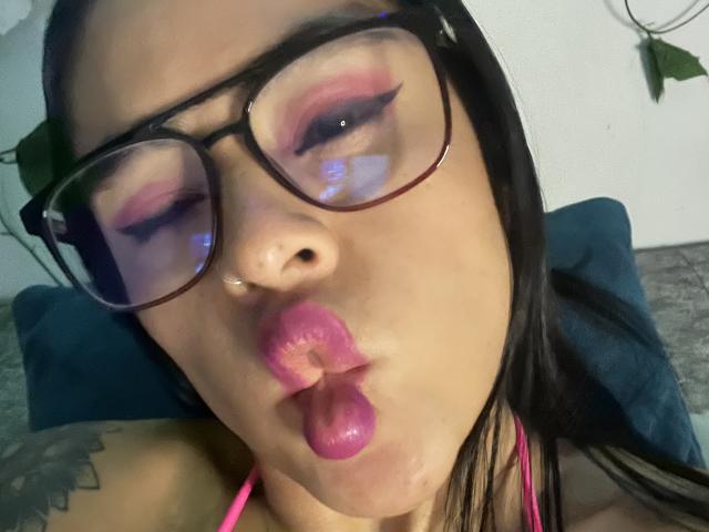 StellaBeckker with pink lipstick, puffy pink-stained lips, and tongue out, wearing glasses, has a tattoo on her shoulder, and is looking at the camera with blue reflections.