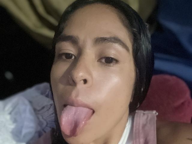 StellaBeckker has tan skin, black hair, and is sticking out her pink tongue with her eyes half-open. She has a small nose piercing and is wearing a pink top.