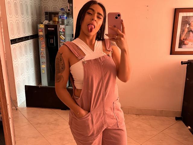 StellaBeckker with pink overalls, white tank top, and hands in pockets, sticking out her tongue, holding a pink phone, tattoo on left arm, in a warmly lit room.