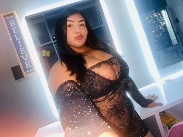 DojaAissha in a black, sheer, lace-up lingerie dress with sparkles, showing her big boobs and curvy ass, standing with a confident, slightly pouty expression.