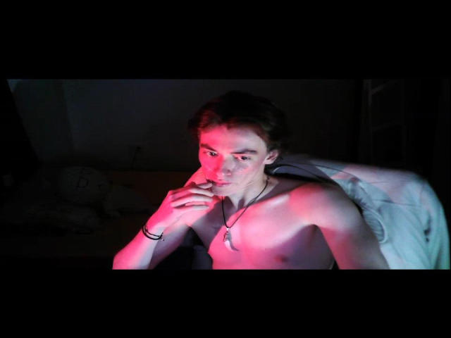 Shirtless CalciferS with pink and blue lighting, right hand near mouth, necklace, black wristband, dark room, slightly smiling, medium build, brown hair, visible chest, subtle shadows.