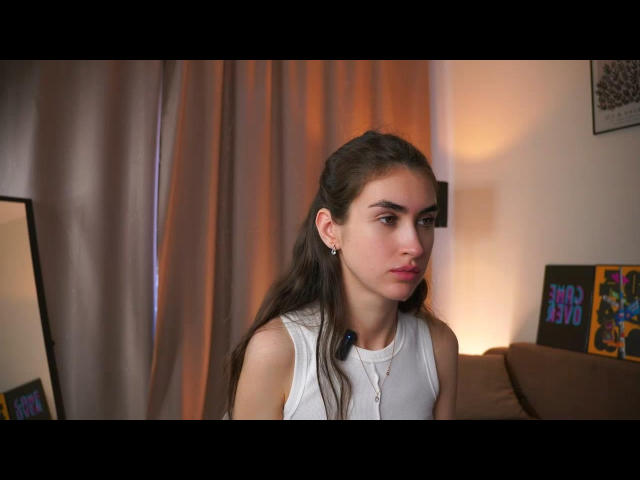 LakitaTigney has long brown hair, wearing a white sleeveless top, looking down with a slightly pouty face, small earrings, and a necklace in a softly lit room.