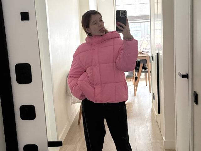 AudryRomie with pink puffy jacket, black pants, taking mirror selfie, tongue out, holding phone, white hallway, wooden floor, black door handles.