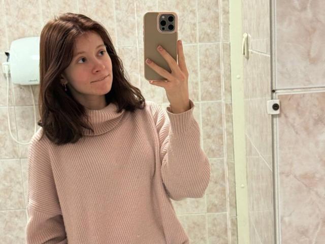 AudryRomie, con cabello castaño medio, un suéter rosa acanalado, sosteniendo un iPhone, tomando una selfie frente al espejo, con una ligera sonrisa, piel pálida, pared con azulejos, un dispositivo blanco montado en la pared, una puerta de vidrio a la derecha..