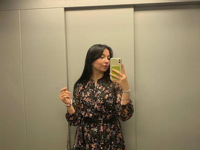 MertieDasinger with long black hair, wearing a floral dress, holding a phone, standing in a room, looking at the mirror, holding a cigarette in her right hand.