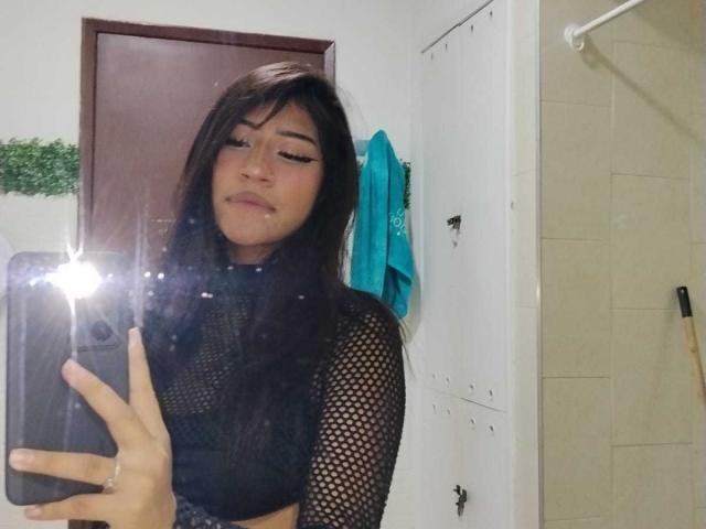 BonnieBond with long black hair, wearing a black mesh top, taking a selfie in a bathroom mirror, showing a subtle smile, with a bright camera flash in the foreground.
