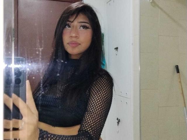 BonnieBond met lang zwart haar, die een zwarte kousenbroektop draagt, houdt een telefoon vast in een spiegelzelfportret, waarbij haar buik en kleine borsten zichtbaar zijn. Ze heeft donkere lipstick en eyeliner..