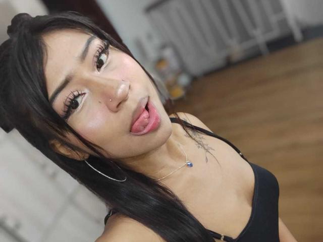 BonnieBond has black hair, a nose ring, and is sticking out her tongue. She's wearing a black top and silver hoop earrings, with a small tattoo on her chest.