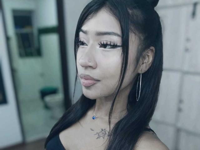 BonnieBond heeft lang zwart haar in een knot, grote wimpers, een neusring, oorringen, een zwarte top, een blauwe ketting en een tatoeage op haar borst. Ze kijkt naar de camera..