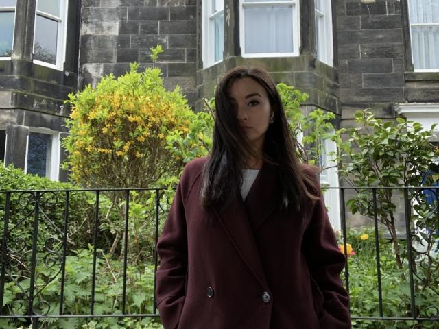 Dionisia, avec ses longs cheveux noirs, portant un manteau couleur bordeaux, se tient devant une clôture en fer noir, des buissons verts et un bâtiment en pierre avec des fenêtres à encadrement blanc..