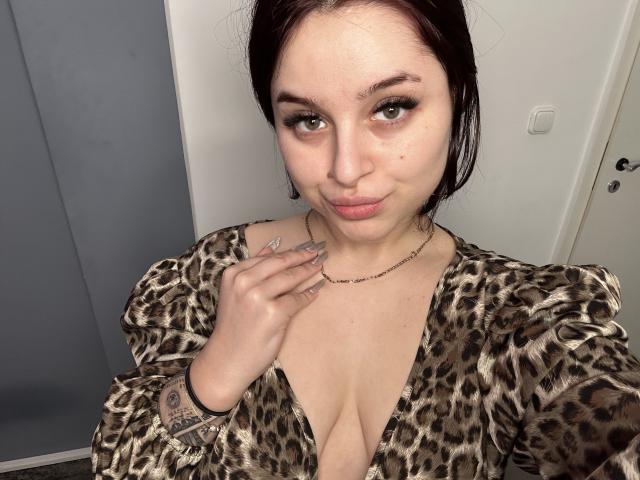 LucieRed with brown hair, green eyes, and a slight pout, wearing a leopard print top with a low neckline, showing her tits. She's touching her necklace with a tattooed hand.